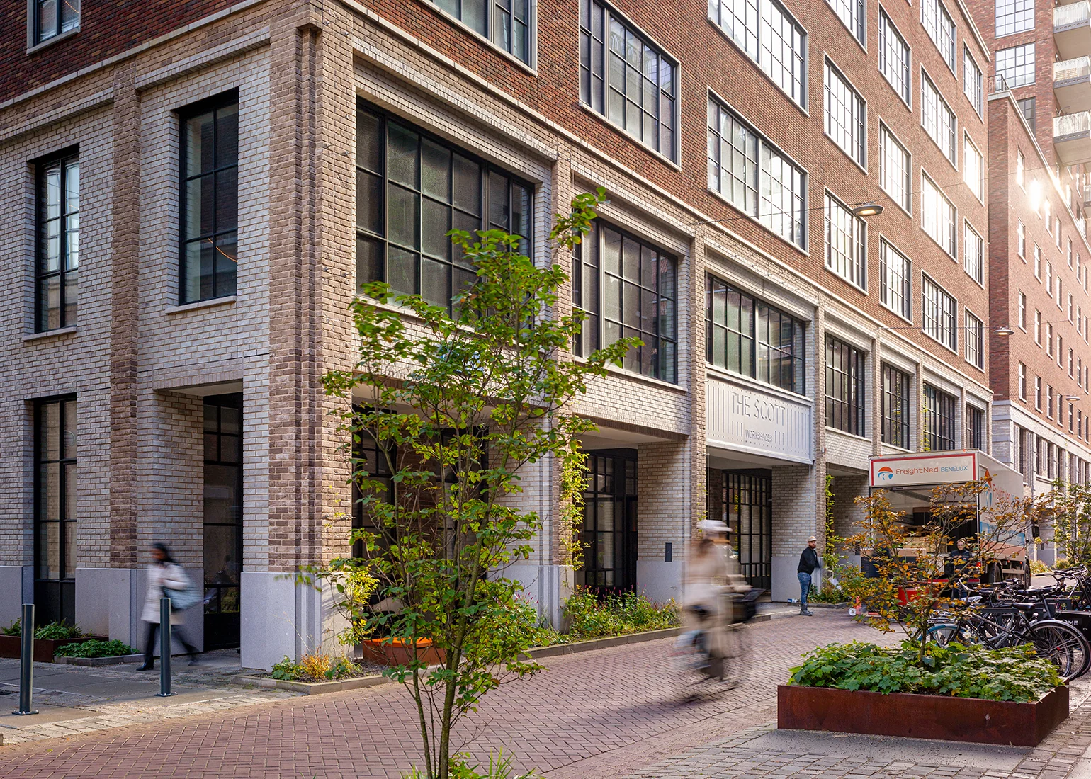 Moderne straat met bakstenen gebouwen aan de Karl Weisbardstraat, met voorbijgangers, fietsen en een foodtruck.